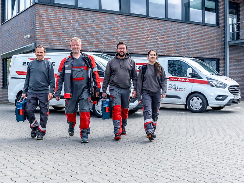 Vier ELMATIC-Mitarbeitende in Arbeitskleidung gehen auf den Betrachter zu, im Hintergrund Firmenwagen